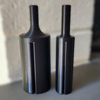 Two black cylindrical metal tool bits with a flared base and narrow shafts, standing upright on a gray surface against a softly blurred light background.