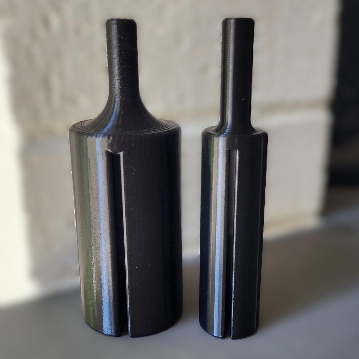 Two black cylindrical metal tool bits with a flared base and narrow shafts, standing upright on a gray surface against a softly blurred light background.