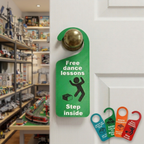 A green novelty door hanger sign with a brass doorknob attachment, hanging on a white door. The sign reads 'Free dance lessons' with a stick figure dancing around a Lego brick and 'Step inside' text.