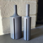 Two matte gray 3D-printed cylindrical bottle-like objects with narrow necks and vertical slit details, standing on a surface against a softly blurred light beige wall.